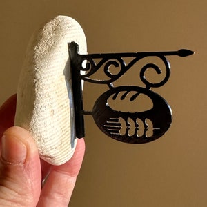 Puede incluir: Una piedra blanca con un letrero de metal negro unido a ella. El letrero tiene la forma de una hogaza de pan con tallos de trigo en la parte inferior.