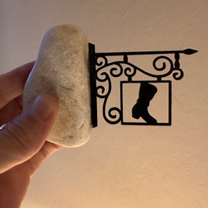 May include: A black metal sign with a boot silhouette in a square frame. The sign is attached to a decorative scrollwork bracket. A light gray rock is in the foreground.