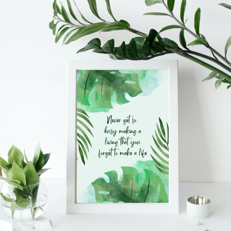 K&ouml;nnte beinhalten: Ein wei&szlig;er gerahmter Druck mit gr&uuml;nem Aquarellhintergrund, der tropische Bl&auml;tter und das Zitat "Werde nie so besch&auml;ftigt mit dem Geldverdienen, dass du vergisst zu leben." zeigt.