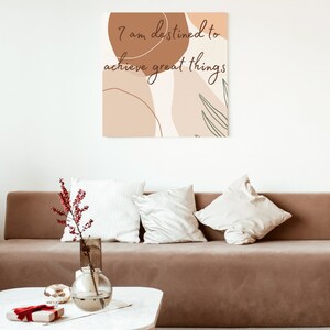 May include: A brown couch with white pillows in a living room with a white wall and a canvas print with the text "I am destined to achieve great things" in brown lettering on a background of abstract shapes in brown and beige.