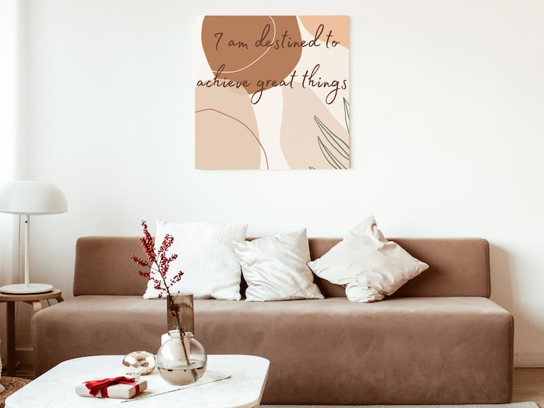May include: A brown couch with white pillows in a living room with a white wall and a canvas print with the text "I am destined to achieve great things" in brown lettering on a background of abstract shapes in brown and beige.