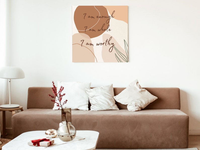 May include: A brown couch with white pillows in a living room setting. A framed print with abstract shapes and the text "I am enough, I am whole, I am worthy" hangs on the wall above the couch.
