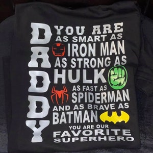 Puede incluir: Una camiseta negra con un gráfico blanco que dice "DADDY" con personajes de superhéroes y el texto "Eres tan inteligente como Iron Man, tan fuerte como Hulk, tan rápido como Spiderman y tan valiente como Batman. Eres nuestro superhéroe favorito."