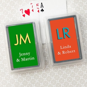 May include: Two personalized playing card decks with green and orange backgrounds. The green deck has the initials "JM" and the text "Jenny & Martin". The orange deck has the initials "LR" and the text "Linda & Robert".