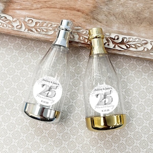 May include: Two clear plastic champagne bottles with silver and gold caps. Each bottle has a round sticker with the text "Ashley & Jake's 25th Anniversary 8.10.25".