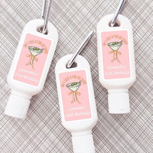 May include: Three small white bottles of sunscreen with pink labels that say "She's just a tiny bit older" with a martini glass graphic and a personalized birthday message. The bottles have a carabiner clip attached.