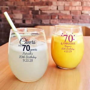 May include: Two clear glass wine glasses with a yellow beverage. One glass has the text "Cheers to 70 years Patrick's 70th Birthday 9.25.28" printed on it. The other glass has the text "70 & FABULOUS Megan's 70th Birthday 4.25.29" printed on it.