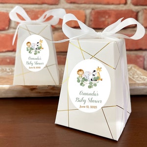 May include: White favor boxes with gold geometric designs and a round sticker featuring a cartoon illustration of a lion, elephant, and giraffe with green leaves. The sticker also includes the text "Amanda's Baby Shower June 12, 2025".