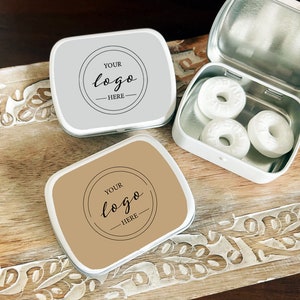 May include: Two small, rectangular metal tins with lids. One tin is silver with a white background and the other is brown with a tan background. Both tins have a circle with the text "YOUR logo HERE" inside. The silver tin is open and contains three white round mints.