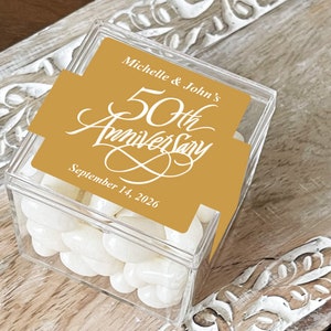 May include: A clear plastic box filled with white candy. The box has a gold label with the text "Michelle & John's 50th Anniversary September 14, 2026".