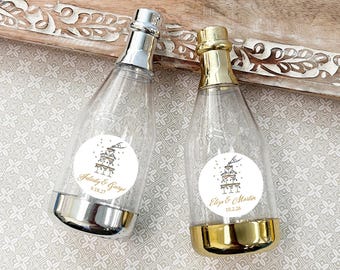 May include: Two clear plastic champagne bottle favors with silver and gold metallic bases and tops. Each bottle has a white circular sticker with a cake design and names and dates. The bottles are on a decorative wooden tray.