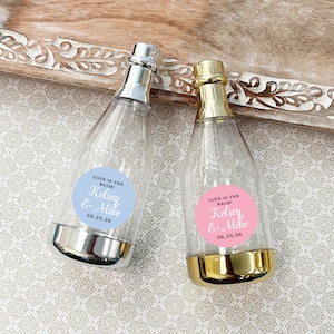 May include: Two clear plastic champagne bottles with silver and gold caps. Each bottle has a round sticker with the text "LOVE IS THE BALM! Kelsey & Mike 06.25.26". The sticker on the left bottle is light blue and the sticker on the right bottle is pink.