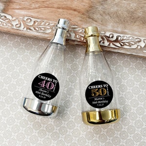 May include: Two clear plastic champagne bottles with gold and silver accents. The bottles have black and white round labels with the text "Cheers to 40 Years" and "Cheers to 50 Years". The labels also include the names "Shayne's 40th Birthday 6.24.26" and "Martin's 50th Birthday 7.12.25".