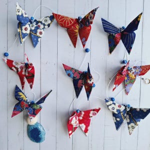 Könnte beinhalten: Eine Girlande aus Origami-Schmetterlingen in Rot, Blau und verschiedenen Mustern. Jeder Schmetterling ist an einem weißen Faden mit blauen Perlen befestigt. Die Schmetterlinge sind mit floralen und geometrischen Designs gestaltet.