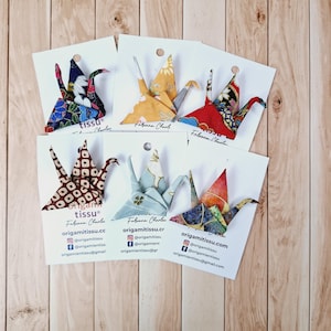 Może przedstawiać: Sześć ozdób w kształcie żurawi origami w różnych kolorach i wzorach, każda umieszczona na białej karcie. Karty zawierają tekst "origamitissu" i dane kontaktowe do mediów społecznościowych.