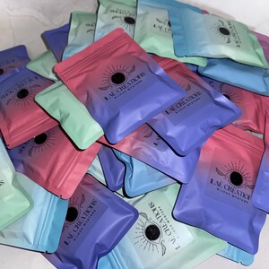 Peut inclure: Une collection de petites pochettes colorées avec un motif répété d'un œil avec des ailes et le texte "LAE CREATIONS BIJOUX MYSTERE".