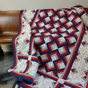 May include: A handmade quilt with a patriotic theme. The quilt features a pattern of red, white, and blue squares and rectangles. The quilt is draped over a wooden bench. The quilt's dimensions are not visible.
