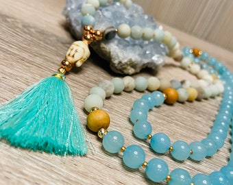 May include: A blue and white beaded necklace with a wooden bead and a small white Buddha charm. The necklace has a turquoise tassel.