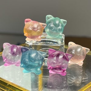 May include: Six small, colorful, translucent cat figurines made of resin. The figurines are in various shades of pink, blue, green, and purple.