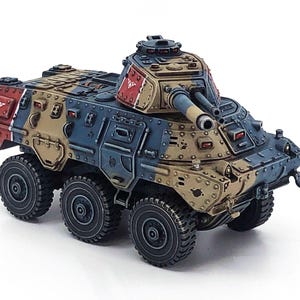 May include: A miniature armored vehicle model with six wheels, painted in a camouflage pattern of tan, blue, and red. The model features a turret with two cannons and is set against a white background.