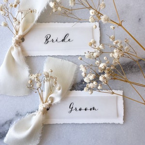 May include: Two white place cards with the words "Bride" and "Groom" written in black calligraphy. Each card is tied with a white ribbon and decorated with a small bouquet of white baby's breath.