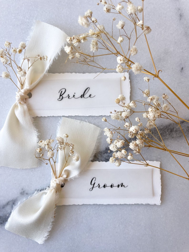 May include: Two white place cards with the words "Bride" and "Groom" written in black calligraphy. Each card is tied with a white ribbon and decorated with a small bouquet of white baby's breath.