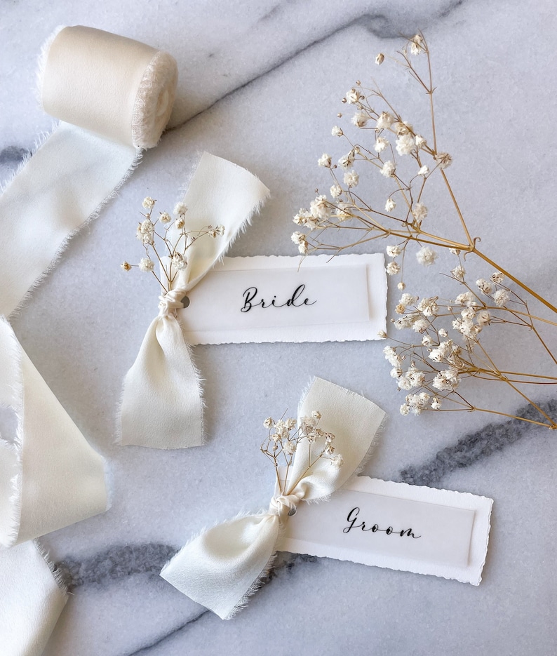 May include: Two white place cards with the words "Bride" and "Groom" written in black calligraphy. Each card is tied with a white ribbon and decorated with a sprig of white baby's breath.