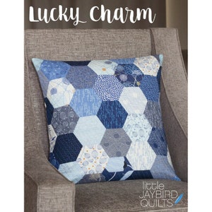May include: A blue and white patchwork pillow with a hexagonal pattern. The pillow is on a gray chair and has the text "Lucky Charm" above it and "little JAYBIRD QUILTS" below it.