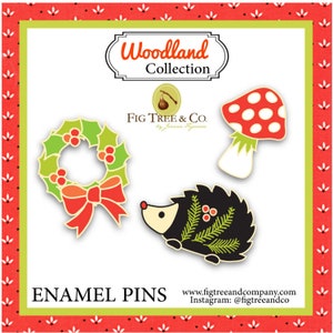 May include: Three enamel pins featuring a red and green holly wreath with a red bow, a black and green hedgehog, and a red and white mushroom with green leaves. The pins are part of the Woodland Collection by Fig Tree & Co.