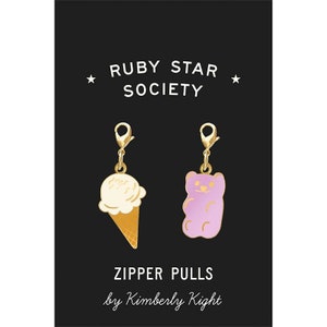 May include: Two gold zipper pulls, one shaped like an ice cream cone and the other like a gummy bear. The ice cream cone is white with a gold cone and the gummy bear is purple. The text "ZIPPER PULLS by Kimberly Kight" is below the zipper pulls.