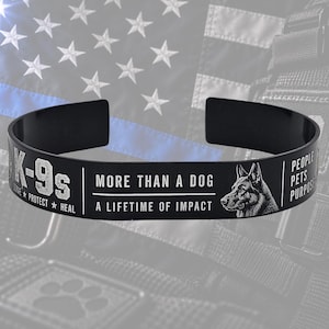 May include: A black metal cuff bracelet featuring white engraved text and a German Shepherd illustration. The text reads K-9s SERVE PROTECT HEAL, MORE THAN A DOG A LIFETIME OF IMPACT, and PEOPLE PETS PURPOSE. The design is centered on a dark background.