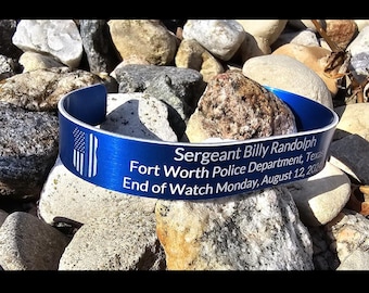Puede incluir: Una pulsera de metal azul con el texto "Sergeant Billy Randolph Fort Worth Police Department, Texas End of Watch Monday, August 12, 200" y un diseño de bandera blanca y azul. La pulsera descansa sobre un lecho de rocas.