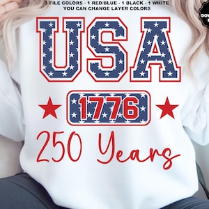 Può includere: Felpa bianca con la scritta "USA" in rosso e blu, con un design a stelle. Sotto, il numero "1776" è visualizzato in uno stile simile, e il testo "250 Years" è scritto in rosso. Il design è patriottico e celebrativo.
