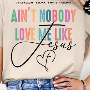 May include: Cream-colored t-shirt with the text "AIN'T NOBODY LOVE ME LIKE JESUS" in colorful block letters. A black cursive design of the word "Jesus" and a heart with a cross is below the text. The shirt also has a small SVG PNG download files logo.