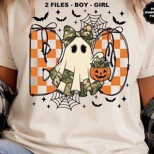 May include: A cream-coloured t-shirt featuring a graphic of a ghost with a camouflage bow and patches, holding an orange jack-o'-lantern bucket. The word BOO is written in orange and white checkers, surrounded by black bats and spider webs.