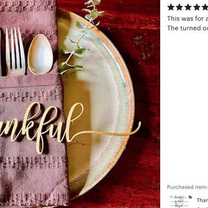 May include: A laser-cut wooden word cutout in cursive script that reads thankful, placed on a dark mauve textured fabric napkin. The napkin holds a silver knife, fork, and spoon, and rests on a round ceramic plate with a light center and rim.