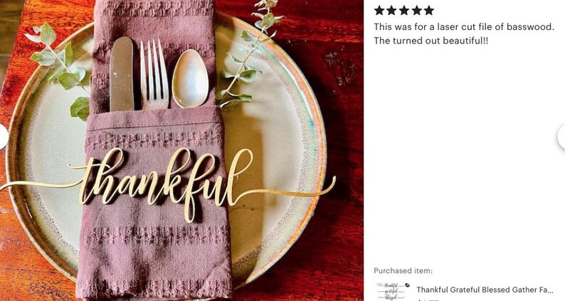 May include: A laser-cut wooden word cutout in cursive script that reads thankful, placed on a dark mauve textured fabric napkin. The napkin holds a silver knife, fork, and spoon, and rests on a round ceramic plate with a light center and rim.