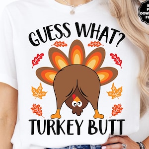 May include: White t-shirt with a humorous Thanksgiving design. The shirt features the text "GUESS WHAT? TURKEY BUTT" in black, with a cartoon turkey illustration in brown, orange, and yellow. Autumn leaves surround the turkey.