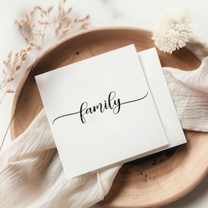 May include: A square white greeting card featuring the word family written in a black cursive script font. The card is positioned on a round wooden tray alongside a piece of white fabric and a small sprig of dried decorative grass.