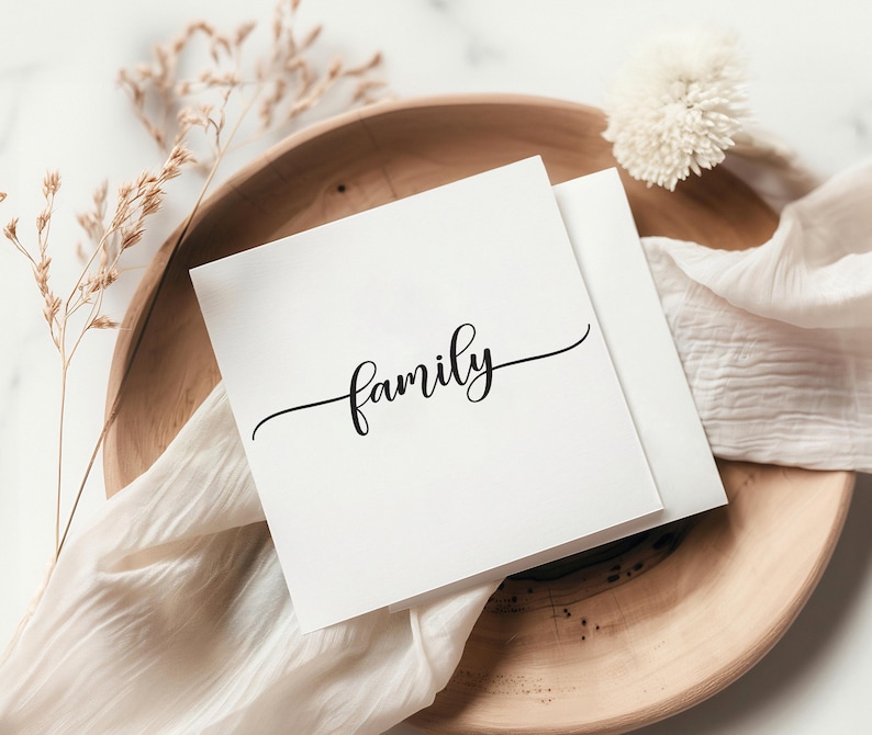 May include: A square white greeting card featuring the word family written in a black cursive script font. The card is positioned on a round wooden tray alongside a piece of white fabric and a small sprig of dried decorative grass.