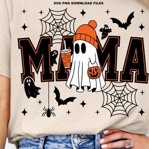 以下が含まれることがあります： ハロウィーンをテーマにしたベージュのTシャツ。デザインは、オレンジ色の輪郭を持つ黒いブロックレターで「MAMA」という言葉を特徴としています。オレンジ色の帽子をかぶり、カボチャと飲み物を持っている幽霊が中央にあり、コウモリ、クモ、クモの巣に囲まれています。