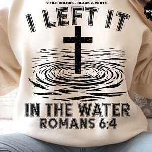 May include: Cream-colored sweatshirt with a black graphic design. The design features a cross above water ripples, with the text "I LEFT IT IN THE WATER ROMANS 6:4". The sweatshirt has a crew neck and long sleeves.