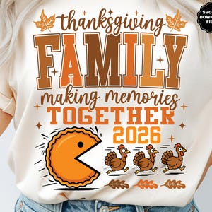 May include: A cream-colored t-shirt featuring a graphic with the text "thanksgiving FAMILY making memories TOGETHER 2026". The design includes an orange pie character chasing three brown turkeys, accented with brown maple leaves and stars.