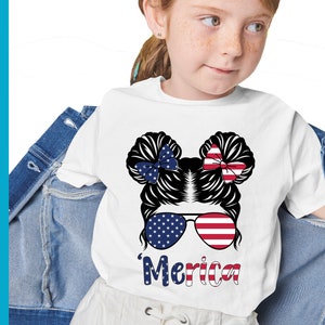 May include: White t-shirt featuring a graphic of a girl with a messy bun, wearing sunglasses with red, white and blue stars and stripes. The text 'Merica' is below the graphic.