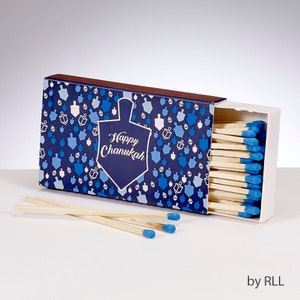 Puede incluir: Una caja de cerillas azul y blanca con una peonza azul y el texto "Happy Chanukah". La caja de cerillas está abierta y muestra cerillas de madera con puntas azules.
