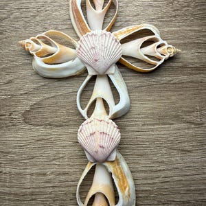 May include: A handcrafted cross made from various seashells, featuring a central design of two scallop shells. The cross is composed of white, tan, and brown shells, arranged on a wooden surface. The shells are intricately shaped, creating a unique religious symbol.