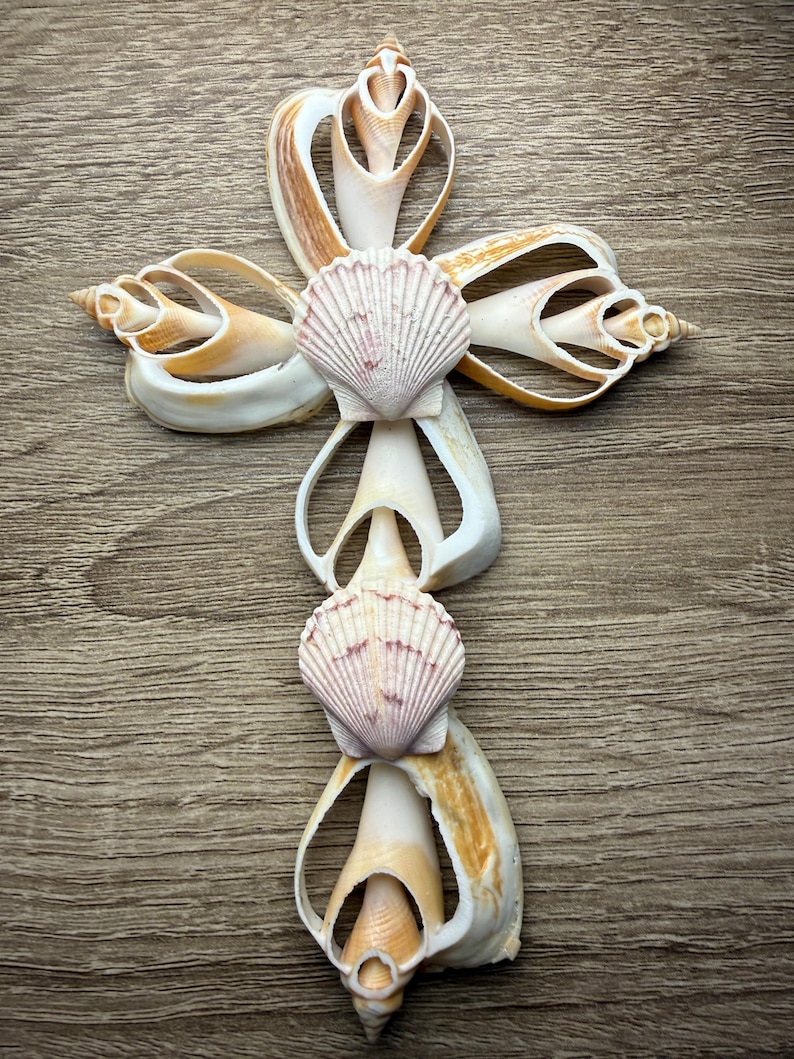 May include: A handcrafted cross made from various seashells, featuring a central design of two scallop shells. The cross is composed of white, tan, and brown shells, arranged on a wooden surface. The shells are intricately shaped, creating a unique religious symbol.