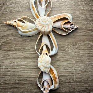 May include: A decorative cross made from various seashells. The cross features a central spiral shell and is adorned with white and tan shells, creating a natural and elegant design. The cross is displayed on a wooden surface.