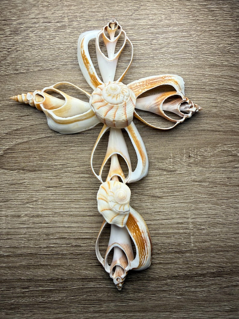 May include: A decorative cross made from various seashells. The cross features a central spiral shell and is adorned with white and tan shells, creating a natural and elegant design. The cross is displayed on a wooden surface.