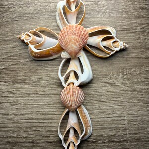 May include: A decorative cross made from various seashells. The cross features a central design of two scallop shells, with other shells forming the arms and base. The shells are primarily cream and tan, with a natural, organic aesthetic.